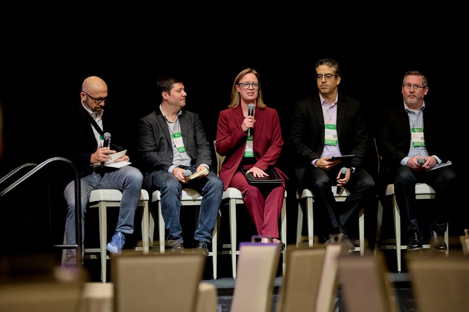 Adrienne Meyer in a maroon suit speaking into a microphone sitting on a stage with two men on eitehr side of her